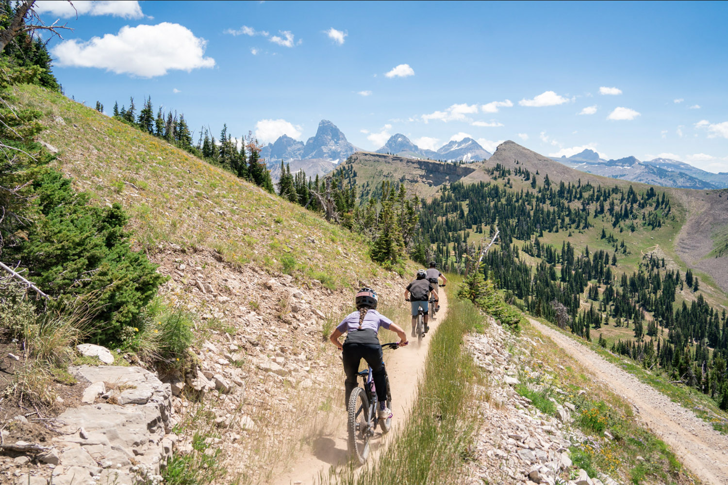 Mountain bikers ride a narrow alpine singletrack trail at Grand Targhee Resort, with sweeping mountain views and evergreen-covered slopes in the background.